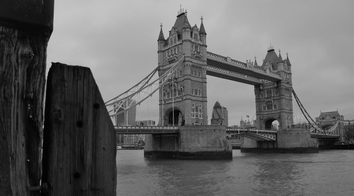 A photo taken from the riverside walkway at Butlers Warehouse, showing the Tower Bridge, Tower of London and other 'towers' of various description, with the Thames flowing by in the middle foreground and an old riverside post in the right foreground 